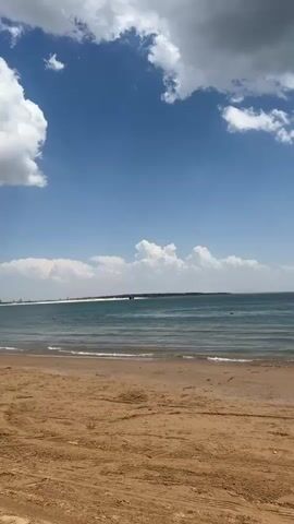 The video shows a sandy beach with the ocean in the background. Several people are standing in the shallow water. Two boats are visible near the shore, one closer to the camera and another further away. The sky is blue with white clouds. The weather appears to be sunny.