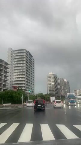 This video captures a view from inside a vehicle driving on a wet, urban street under a heavy, cloudy sky. The road is slick with rain, and several cars are visible, including a black SUV in the foreground. On either side of the street, modern, tall residential buildings dominate the skyline. Lush green trees and vegetation line the sidewalks and surround the base of the buildings. Traffic lights, including a green one, are visible. The overall mood is somber due to the overcast weather. There is no visible text in the frame.