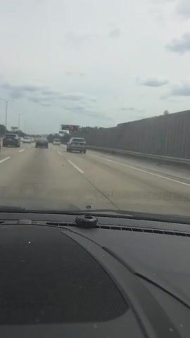 The video shows a first-person view from a car driving on a highway. The sky is overcast with clouds. Several cars are on the highway ahead and to the left. To the right, there is a concrete barrier with a fence or trees behind it. In the distance, a large electronic billboard is visible. The billboard displays text which is partially obscured but appears to read "TIME TO 1:00 I-94/IL 390". The car has a black dashboard with a small black circular object in the center. The windshield has some smudges or water marks. The road appears to be wet, suggesting it may have rained recently or is still raining lightly.