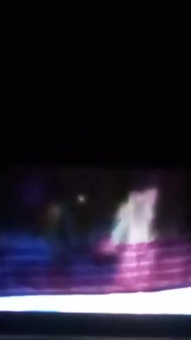 The video is a blurry, dark, low-resolution recording of a group of people in what appears to be a performance or presentation setting. The individuals are largely indistinct, but some are wearing brightly colored clothing, including yellow and pink. The background is dark and indistinct, with some faint lights visible. The people appear to be standing or sitting on raised platforms, and there are horizontal lines that suggest the presence of seating or tiered staging. The overall impression is of a stage production, possibly a children's show or a concert, given the bright colors and the presence of multiple figures.