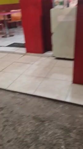 Here is a detailed description of the video:

The video appears to be taken outside of a Dr. Chicken restaurant. The camera is pointed toward the front counter of the restaurant, showcasing its branding. A large, round logo featuring the words "Dr. Chicken" in white and yellow against a red background is prominently displayed. 

The counter itself is red and white, with a glass display case on top. Inside the display case, one can see what appears to be fried chicken, fries, and buns. Two women wearing hijabs are visible behind the counter. One is standing further back, possibly taking an order or doing some work. The other woman is closer to the camera, mostly obscured by the counter.

The flooring in front of the counter is composed of light-colored tiles. The ground in front of the tiles appears to be asphalt. The lighting in the video is somewhat dim, possibly indicating it's evening or nighttime.