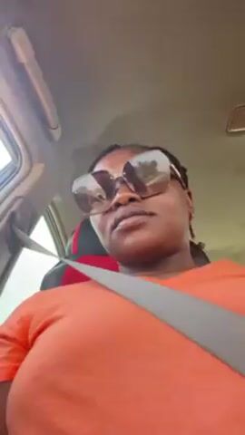 A woman wearing an orange shirt and sunglasses is sitting in a car. She is wearing a seatbelt. The car has a red and black seat cover. The woman has braids. The car's interior is visible. The camera is pointing up at the woman.