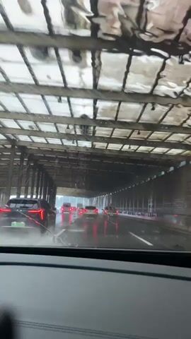The video shows a first-person perspective from inside a vehicle driving through a tunnel on a rainy day. The windshield is wet with raindrops, obscuring the view slightly. Several cars are ahead, their taillights visible, indicating traffic. The tunnel has a metal and wood-like ceiling structure with visible beams and panels, and the walls appear to be concrete. The road is wet and reflects the lights of the vehicles and the tunnel's illumination. The overall atmosphere is dim and wet, typical of driving through a tunnel in bad weather.