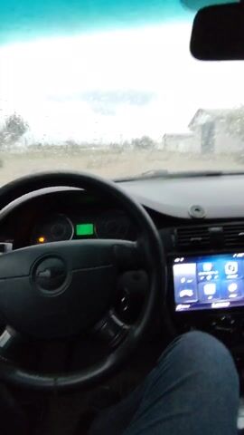 The video shows the inside of a car on a rainy day. The windshield wipers are activated and clear the rain from the windshield. The driver's hand is visible on the steering wheel, and they turn on the windshield wipers. A digital display in the center console shows various app icons. The car appears to be parked on a dirt road near some buildings. The weather is cloudy and overcast.