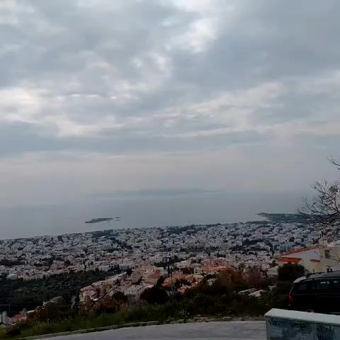 The video shows a cityscape with a sea view under a cloudy sky. The city is densely populated with buildings, mostly white with red roofs. The sea is calm, with a few small islands visible in the distance. In the foreground, there is a road with a small grassy area and some vegetation. A black vehicle and a graffiti-covered container are visible on the right side of the frame. A dog barks in the background.
