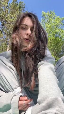 A young woman is featured in a short video clip, sitting outdoors on what appears to be a mat or blanket on the ground. She is dressed in a grey hooded sweatshirt and matching grey sweatpants, with white socks visible. She wears a ring on her right hand. The setting is a natural, wooded area with tall trees and green foliage, under a clear blue sky, suggesting it is daytime and sunny. Throughout the video, she shifts her posture and facial expressions, sometimes looking directly at the camera, sometimes looking away or down. Her long, dark hair moves as she changes her head position. The video has upbeat background music. The location context provided indicates this is in Слизнево, Russia.