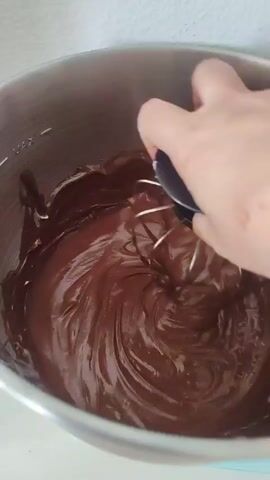 A close-up shot shows a hand holding a black and silver electric hand mixer with white beaters, stirring a thick, dark brown chocolate mixture in a stainless steel mixing bowl. The beaters are partially submerged in the chocolate, creating swirls and waves. The hand appears to have some red smudges on it. The mixing bowl is on a surface that appears to be light colored. The background is a plain, light-colored wall.