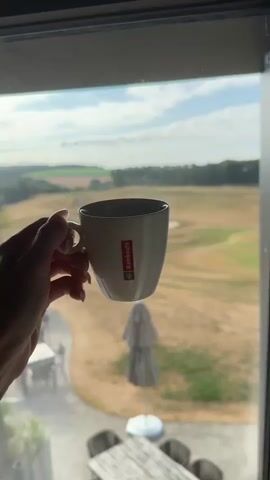 A hand holds a white coffee mug with a red logo in the foreground, obscuring part of the view out a window. Through the window, a wide shot of a dry, brown golf course is visible. The golf course has rolling hills, a paved path winding through it, sand traps, and a pond. In the middle ground, there are several outdoor tables and chairs, and folded umbrellas. The background shows a line of trees and a field under a partly cloudy sky. The overall impression is a calm, leisurely morning scene.