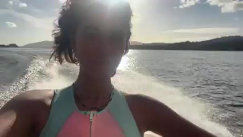 A woman is seen in a selfie-style video taken on a boat moving quickly across a body of water. The sun is behind her, causing lens flare and obscuring some details. The woman has dark hair and is wearing a light-colored wetsuit with a pink panel on the front. She is looking at the camera with a slight smile. The boat is leaving a wake of white water behind it. In the background, there are landmasses, possibly hills or mountains, and a partly cloudy sky.