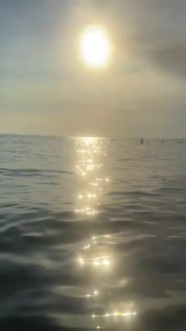 The video shows a serene view of the ocean with the sun shining brightly and reflecting on the water. There are a few distant figures visible in the water, possibly people swimming or standing. The sky is a soft blue with some clouds. The water appears calm with gentle ripples. The sun is positioned high in the sky, casting a shimmering path of light across the surface of the ocean. There are some dark, vertical objects in the distance, which could be buoys or markers. The overall atmosphere is peaceful and tranquil.