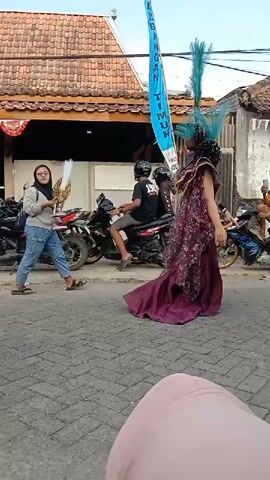 The video shows a street scene with several people and motorcycles. On the left, a person in a red costume with a feathered headdress is partially visible. In the center, a person in a gray hoodie and blue jeans is interacting with a person on a motorcycle. The person on the motorcycle is wearing a helmet and a black shirt. To the right, two individuals are walking in elaborate costumes. The first person on the right is wearing a dark, long, flowing dress with intricate patterns and a large, black, feathered headdress. The second person, further to the right and slightly behind, is wearing a red outfit with gold accents and a tall, feathered headdress. Several motorcycles are parked on the side of the street. In the background, there is a building with a tiled roof and some decorative banners. The ground is paved with interlocking bricks. The sky is visible and appears to be daytime.