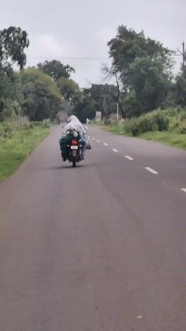 Two people are riding a motorcycle on a paved road. The person in front is driving, and the person behind is a passenger. Both are carrying bags. The scene appears to be in a rural area with trees and greenery lining the road. The sky is overcast.