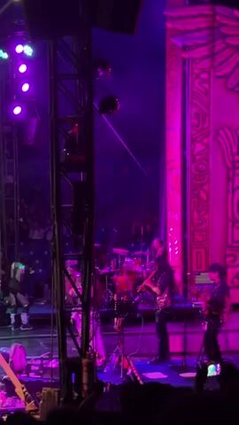 The video shows a performance taking place on a stage, illuminated by purple and pink lights. In the background, a large, circular cage, resembling a globe of death, is visible. Several performers, dressed in costumes that appear to be part of an act, are spread across the stage. Some are positioned near the globe, while others are further forward. There are also indications of a live band, with instruments like drums and possibly a trumpet visible on the right side of the stage. The audience is present, with hands raised in anticipation or applause in the foreground. The atmosphere is energetic and typical of a live entertainment event.