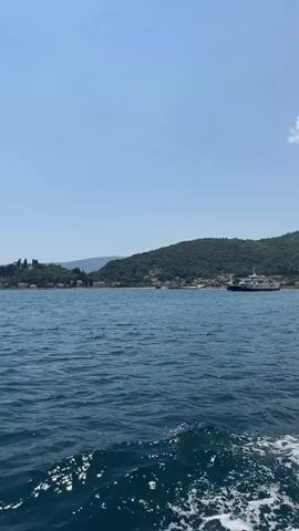 This media file captures a scenic view of the sea near Đurići, Montenegro. The foreground is dominated by the choppy, deep blue water, with sunlight glinting off the surface and creating white foam trails from a boat moving out of frame. In the middle ground and background, a lush, green, hilly coastline stretches across the horizon. Dotted along the coastline are various buildings, including what appear to be houses and a church with a steeple, suggesting a populated area or small town nestled into the landscape. Further out on the water, a ferry or a larger boat is visible, hinting at maritime activity.

The sky is a clear, bright blue, indicating fair weather and likely a sunny day. The overall impression is one of a beautiful, tranquil coastal scene, possibly during the daytime, given the clear visibility and bright sunlight. The absence of people in the immediate frame and the vastness of the sea and coastline evoke a sense of peacefulness and natural beauty. There is no visible text within the media file.