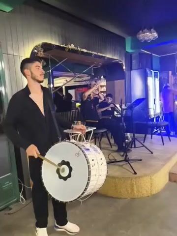 A young man with dark hair and a beard, wearing a black shirt and black pants, stands in the foreground playing a large white drum with a wooden mallet. The drum is suspended by a strap. In the background, on a raised stage, two other men are playing instruments. One man is playing a stringed instrument, possibly a violin or a lute, and the other is playing a wind instrument, possibly a clarinet or a zurna. They are seated on chairs and have microphones in front of them. The room appears to be a bar or restaurant, with a bar area visible behind the musicians and blue lighting in the background. There are speakers and stage lighting present. The overall atmosphere suggests a live musical performance.