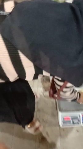 A person is standing in a market. The person is wearing a black and beige striped sweater, black pants, and beige sandals. They are holding two green melons. In the background, there is a scale and shelves filled with various items. The floor is tiled. The video is shaky and has a low resolution.