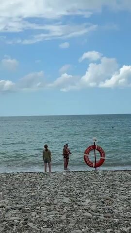 The video shows a beach scene with a person on the left and a person with a child in the water on the right. There is a life preserver on a pole to the right of the people. The water is choppy and the sky is cloudy. The person on the left is facing away from the camera. The person holding the child is facing the water. The child is facing the camera. The life preserver is orange and white with text on it. The beach is covered in pebbles.