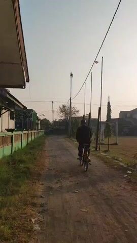 A person rides a bicycle on a dirt path. The person is wearing dark clothing and is moving away from the camera. The path is lined with grass and a green fence on the left, and a sports field with soccer goals and tall poles on the right. The sky is clear and light, indicating either sunrise or sunset. The environment appears to be outdoors in a rural or semi-rural area, possibly near a school or sports facility.