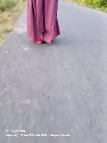 A person wearing a pink skirt and a black headscarf walks away from the camera down a paved road. The person's back is to the camera. The road is surrounded by green foliage and trees. The sky is overcast. A timestamp in the bottom left corner reads "OPPO A6 Pro fataelnurmy 25 Juni 2026 pukul 06.25 Kabupaten Bireuen."