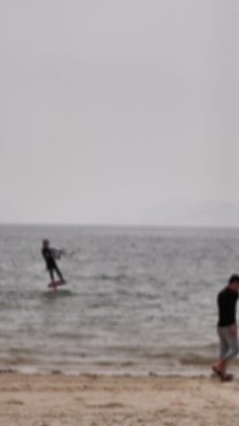 On an overcast day in Bai Chay, Vietnam, a man in a dark wetsuit is kitesurfing on the water near a sandy beach. He stands on a board, holding onto a control bar connected to a kite that is largely out of frame above him. The water is choppy, reflecting the muted light from the grey sky.

Another man stands on the light-colored sandy shore, observing the kitesurfer. He raises a cell phone to record or photograph the activity. In the distant background, faint landmasses and horizontal power lines stretching across the water are visible. The scene depicts a leisure activity on a calm, cloudy day.