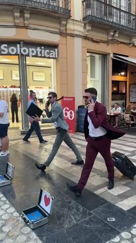 Two men walk across a pedestrian street, one in a gray suit and the other in a burgundy suit, both talking on cell phones. The man in the gray suit carries a black briefcase. The man in the burgundy suit pulls a black rolling suitcase. In the foreground, two open silver briefcases are on the ground, one with a red heart graphic on the lid and the other containing coins. In the background, people walk past a Soloptical store, and some people are seated at outdoor tables of a cafe.