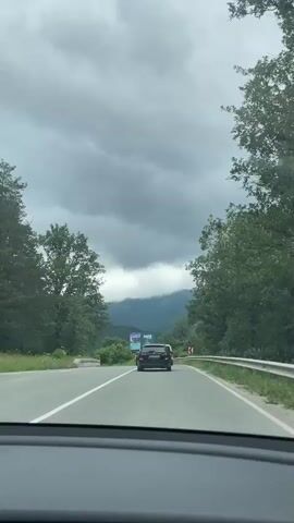 The video shows a view from inside a car traveling on a road. There are three cars in front of the camera's vehicle, moving in the same direction. The road is paved with white lane markings. On the right side of the road, there is a guardrail and a billboard advertising a hotel or resort called "SANTÉ" with an image of a swimming pool. The left side of the road is bordered by trees and vegetation. In the distance, there are mountains covered with trees, and the sky is overcast with dark clouds. The overall atmosphere suggests a cloudy or potentially rainy day in a mountainous, rural area. The video is short and does not contain any audio.