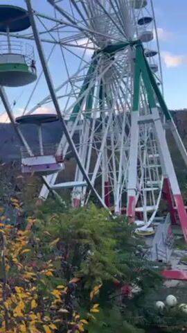 The camera pans up an old Ferris wheel with red, white, and green supports, with a view of trees and distant hills in the background.  The sky is partly cloudy.  The Ferris wheel is not operating.