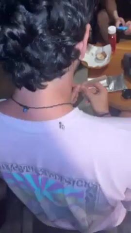 Here is a detailed description of the video:
The video shows the back of a person with curly black hair, wearing a light-colored t-shirt with a graphic design and a black necklace with a blue pendant. The person is sitting at a table and appears to be rolling a cigarette or joint. There are other people in the background, along with items on the table like food, a ketchup bottle, and what looks like aluminum foil. The lighting is somewhat dim.