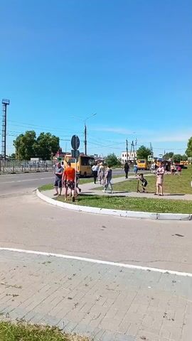 The video displays an outdoor scene on a sunny day with a clear blue sky. The foreground features a paved road leading to an intersection with a circular grassy median. Multiple pedestrians, including adults and children, are visible walking on the sidewalks surrounding the median and crossing the road. On the left, people walk along a paved path next to a street. On the right, individuals are walking and standing on a sidewalk adjacent to the grassy median. In the background, a street with two yellow buses is visible, along with distant buildings, power lines, and trees. A lamppost stands prominently on the right side of the frame, and street signs are present near the intersection.