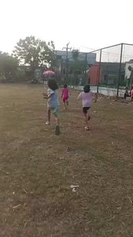 Three young girls are running across a grassy field. The girl in the foreground is wearing a light blue t-shirt and light green shorts. She is running away from the camera. The second girl, in the middle, is wearing a pink t-shirt and black shorts. The third girl, in the background, is wearing a pink dress and is slightly ahead of the second girl. There is a pink umbrella visible near the second girl. The field is mostly brown grass with some scattered leaves. In the background, there are trees, buildings, and a fence. The sky is bright and appears to be either sunrise or sunset. The overall impression is of children playing outdoors.