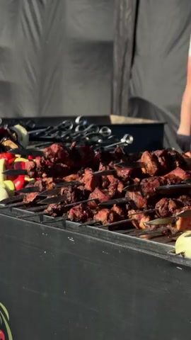 The video shows an outdoor food stall with several skewers of meat and vegetables grilling over an open fire. In the background, there are people moving around and a few tents set up, suggesting a market or festival. The stall itself is decorated with a colorful floral pattern. The focus of the video is on the food being prepared, with close-ups of the grilling skewers and the raw ingredients. The overall impression is one of a lively and bustling outdoor dining experience.