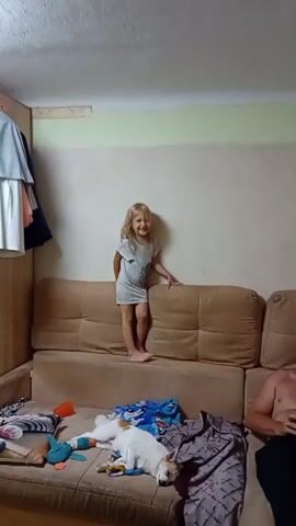 A young girl with blonde hair and a light blue dress stands on the back of a brown couch. Her mouth is open, and she appears to be laughing or speaking. Her hands are resting on the back cushions of the couch. A small dog is lying on the couch in front of her, appearing to be asleep. The dog is white with brown markings on its head and back. Various items are scattered on the couch, including a blue blanket, a toy, and some clothing. The room appears to be a living space, with a closet on the left side of the frame and a wall with pale green and beige paint behind the girl. A person's arm is visible on the right side of the frame.