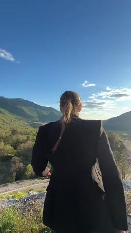 A woman with a ponytail wearing a black blazer and jeans stands on a hilltop overlooking a valley on a sunny day. The sky is blue with a few scattered clouds.  She raises her arms in the air, facing the valley.  Mountains are visible in the background. A road winds through the valley below. Music plays.