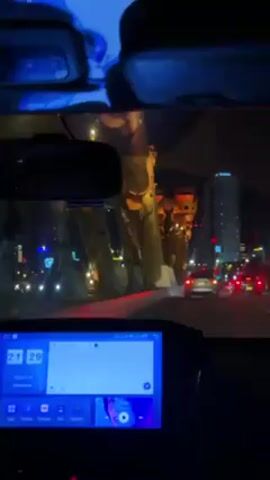 The view from inside a car driving at night through a city. The car's dashboard is visible, displaying a digital clock that reads 21:29 and a date of 2024/03/12. The car's interior is dark, but the ambient city light illuminates the dashboard screen. Through the windshield, one can see the cityscape at night, including a tall, ornate, orange-lit bridge structure. There are other cars on the road, indicated by their headlights and tail lights. In the background, a tall building is visible, contributing to the urban skyline. The rear-view mirror also reflects the scene behind the car, although the details are obscured by the darkness. The overall atmosphere is that of night-time urban driving.