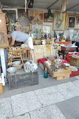 The video shows a person reaching into a stall filled with various antiques and bric-a-brac. The stall is set up outdoors, possibly at a market, with items displayed on shelves, tables, and the ground. The items include paintings, lamps, figurines, crockery, and old luggage. The person is wearing a light-colored shirt and shorts and is bent over, seemingly examining something within the stall. The overall impression is of a cluttered but organized collection of vintage items.