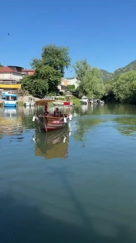 A motorboat with two people on board moves toward the camera on a river. The river is surrounded by trees and buildings. The sky is clear and blue.