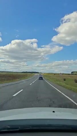 The video shows a point-of-view shot from inside a car driving on a two-lane highway. The sky is blue with scattered white clouds. On either side of the road are fields of grass and crops, with some trees and buildings visible in the distance. A single car is in front of the vehicle, and a road sign indicating a curve is visible on the right side of the road. The camera is positioned behind the windshield, and the dashboard is partially visible at the bottom of the frame.