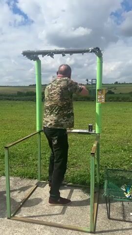 A man stands in a shooting stall and fires a shotgun. The man is wearing a camouflage shirt and black pants. He is bald and has a beard. He is holding the shotgun with both hands and aiming it at a target that is not visible in the frame. The shooting stall is made of metal and has green posts. There is a grassy field behind the stall, and a cloudy sky above. A basket of colorful shotgun shells is visible to the right of the stall.