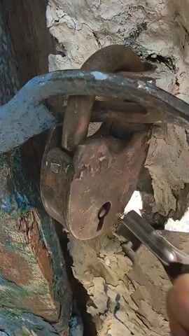 A hand is inserting a key into an old, rusty padlock that is securing a metal shackle to a textured, light-colored surface. The padlock is partially obscured by the shackle and the surrounding material. The key is being turned, and the padlock appears to be opening. The lighting suggests an outdoor setting.