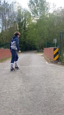 A person wearing a two-toned hooded sweatshirt and sweatpants is standing on a paved road, then steps up onto a low brick wall with yellow-and-black stripes on the side, stepping over the green metal fence and into the grassy area behind it. The person is facing away from the camera. There are trees on either side of the road. The sky is overcast.