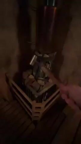 A person is pouring water onto a stone-filled heater in a sauna. The heater is encased in a metal cage and surrounded by a wooden railing. Above the heater is a copper-colored cylinder. The sauna has a wooden floor and walls. The video is dark.