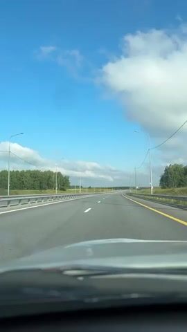 The video shows a view from the windshield of a car driving on a highway. The road is asphalt with white and yellow lane markings. There are guardrails on both sides of the road. Trees and fields are visible beyond the guardrails. The sky is blue with scattered white clouds. Streetlights are visible along the side of the road.