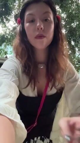A young woman with brown wavy hair, adorned with red hair accessories, stands outdoors in a gravelly courtyard. She wears a white off-the-shoulder blouse with wide sleeves, a black corset-like top, a black and white checkered skirt, and a bright pink crossbody purse. The woman is posing and dancing, moving her arms and body. Trees and greenery are visible in the background, along with a dark-colored door and decorative statues. The lighting suggests an overcast day or late afternoon.
