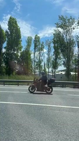 The video shows a motorcycle traveling on a highway. The rider is wearing a helmet and dark clothing. The motorcycle is dark in color with a storage case on the back. The road is gray and has white lines marking the lanes. Trees and grass are visible on the side of the road. The sky is blue with scattered white clouds. There are also some buildings visible in the background. The camera appears to be moving alongside the motorcycle. A sign indicating various services is visible at the end of the video.