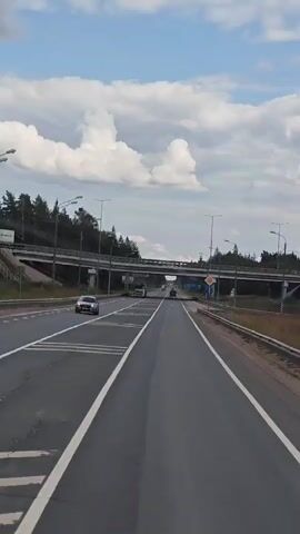 The video shows a view from a vehicle traveling on a highway. The highway has multiple lanes and is bordered by a guardrail on the right. There are trees and a bridge over the highway in the distance. Several trucks and cars are visible on the road. The sky is blue with white clouds.