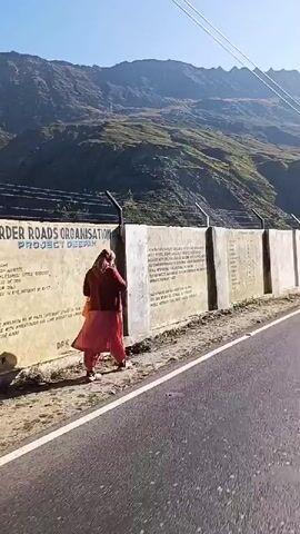 The video shows a first-person perspective of a motorcycle ride on a paved road. The road curves to the left, with a concrete wall on the right side. The wall has some writing on it, including a list of distances and the phrase "HEAVEN OR HELL IS ANOTHER MAN'S CHOICE & MOTIVES". Behind the wall, a steep, rocky mountain slope is visible, covered in vegetation. Power lines run overhead. The sky is clear and blue, with the sun shining brightly. In the foreground, the back of a motorcycle helmet is visible, with the brand name "GLIDERS" clearly displayed. The overall impression is a scenic journey through a mountainous region.