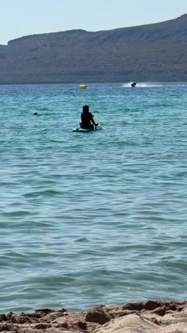 A person sits on a paddleboard in the middle of the ocean, holding a paddle. In the distance, a jet ski speeds across the water. The sky is clear and the sun is shining. There is a sandy beach in the foreground and a hilly, vegetated landscape in the background. The water is clear and turquoise, with visible waves.