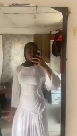A person wearing a black hijab and white dress is taking a selfie in a mirror. The mirror is framed and slightly tilted. The background includes a patterned wall, a wooden ceiling, and various items like clothes hanging behind the person. The person is holding a smartphone in their right hand, using it to capture the image.