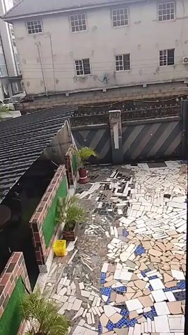 The video shows an outdoor patio area, likely viewed from an elevated position. The patio has a floor made up of broken tiles in various shades of white, blue, and beige, creating a mosaic-like appearance. A low wall built with brick and green artificial turf runs along one side of the patio. Two potted plants are placed along this wall, with a yellow plastic container nearby. Another, taller potted plant is situated near a gate. The gate is painted with a gray and black chevron pattern. A corrugated metal roof slopes downwards behind the wall, and a multi-story building with numerous windows is visible in the background. The video appears to be taken on a bright day, and the overall impression is one of an unfinished or unconventional outdoor space. In the beginning, there seems to be sound that could be a siren.