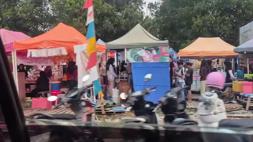 The video depicts an outdoor market scene, viewed from inside a vehicle. Numerous tents are set up, displaying various goods and services. People are seen browsing and interacting at the stalls. Motorcycles are parked in the foreground. The environment appears to be a street or roadside market with trees visible in the background. The overall impression is one of a bustling commercial area.
